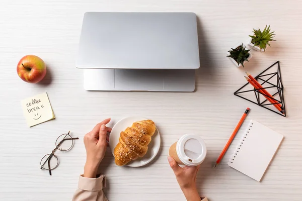 Tiro recortado de mãos femininas com copo de café descartável e croissant, laptop e material de escritório na mesa — Fotografia de Stock