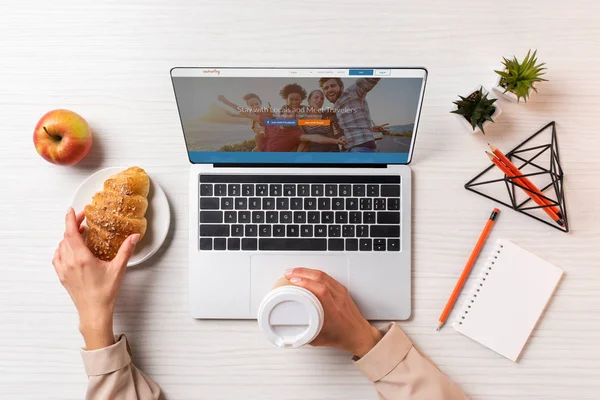 Colpo ritagliato di donna d'affari in possesso di tazza di caffè usa e getta e croissant durante l'utilizzo del computer portatile con sito web couchsurfing — Foto stock