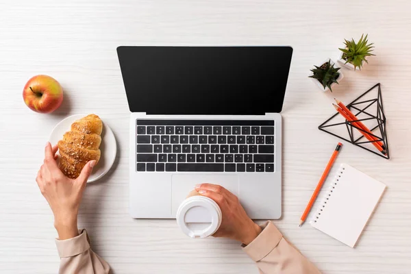 Cropped shot of businesswoman holding disposable coffee cup and croissant while using laptop with blank screen — Stock Photo