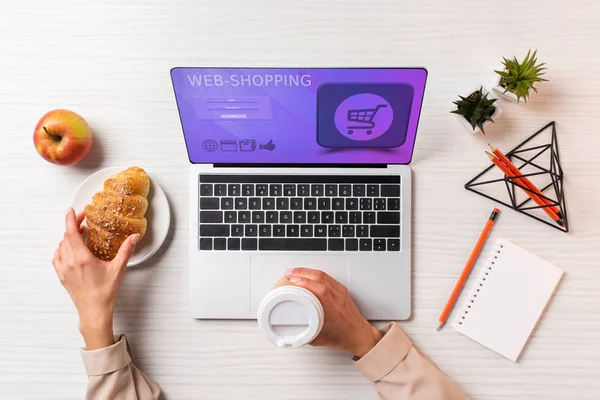 Recortado tiro de mujer de negocios sosteniendo taza de café desechable y croissant mientras se utiliza el ordenador portátil con aplicación de compras web - foto de stock