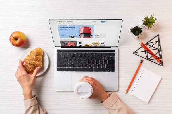 Cropped shot of businesswoman holding disposable coffee cup and croissant while using laptop with ebay website — Stock Photo