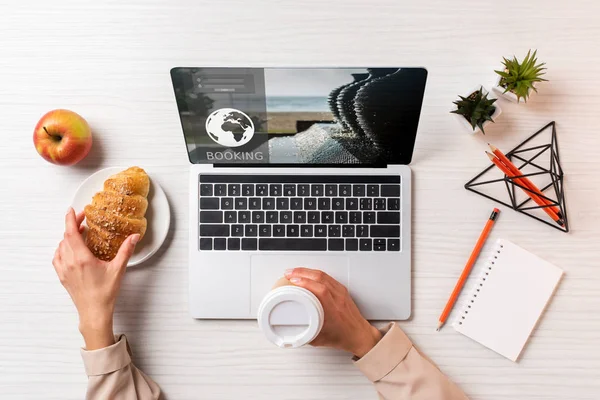 Cropped shot of businesswoman holding disposable coffee cup and croissant while using laptop with booking website — Stock Photo