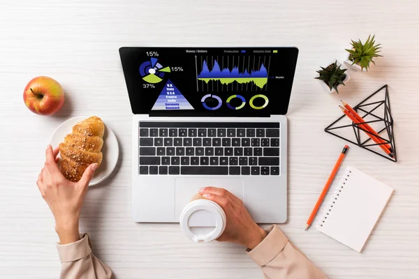 Cropped shot of businesswoman holding disposable coffee cup and croissant while using laptop with business charts — Stock Photo