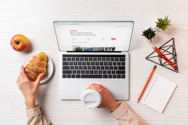 Cropped shot of businesswoman holding disposable coffee cup and croissant while using laptop with airbnb website — Stock Photo