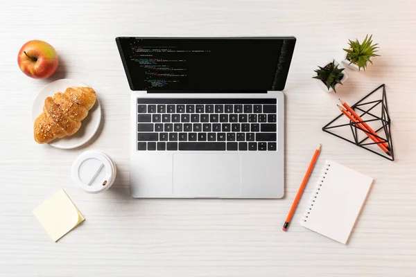 Vista superior do laptop com código html na tela, xícara de café descartável, maçã e croissant na mesa — Fotografia de Stock