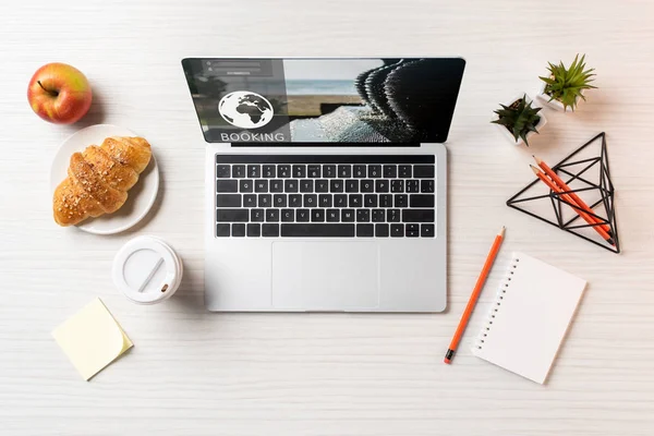 Vue du haut de l'ordinateur portable avec site de réservation, croissant, pomme et café pour aller sur la table de bureau — Photo de stock