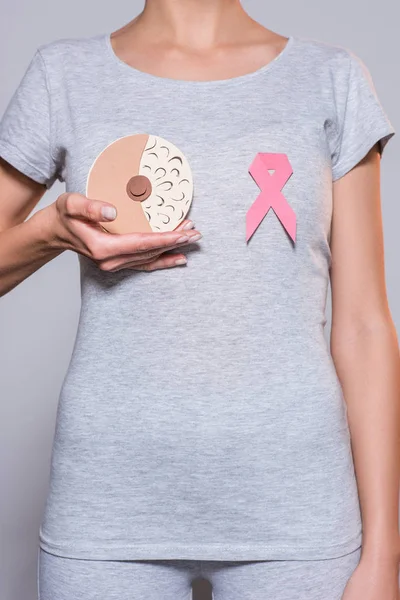 Partial view of woman with paper made female breast and cancer awareness pink ribbon on grey background — Stock Photo