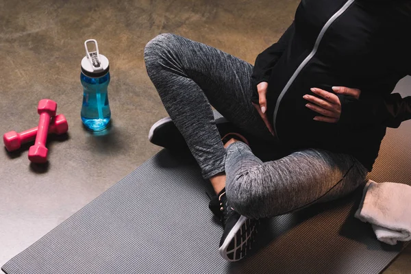 Vista recortada de la mujer deportiva embarazada sentada en la alfombra deportiva y la botella de deporte al lado — Stock Photo