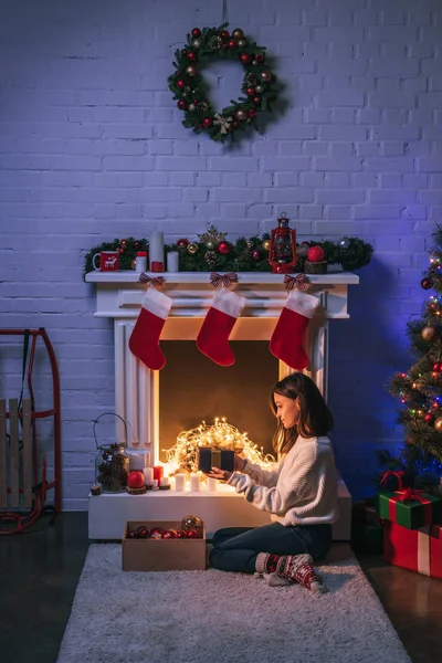 Schöne Frau hält Geschenk und sitzt in der Nähe von Weihnachten dekorierten Kamin und Weihnachtsbaum — Stockfoto