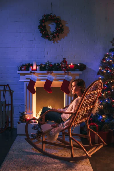 Mulher feliz sentada na cadeira de balanço perto de lareira decorada e árvore de Natal — Fotografia de Stock