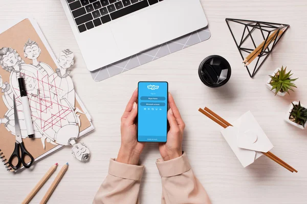 Office desk with laptop and woman hands holding smartphone with skype app on screen, flat lay — Stock Photo