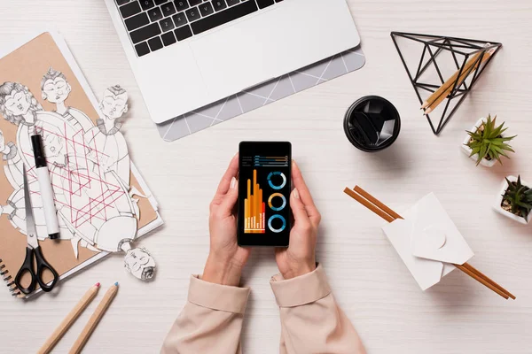 Escritorio de la oficina con el ordenador portátil y las manos de la mujer sosteniendo el teléfono inteligente con gráfico en la pantalla, disposición plana - foto de stock