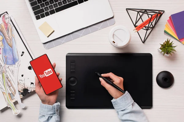 Cropped view of designer using graphics tablet, pen and smartphone with youtube app on screen, flat lay — Stock Photo