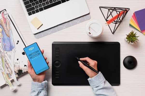 Cropped view of office desk with designer using graphics tablet, pen and smartphone with skype app on screen, flat lay — Stock Photo