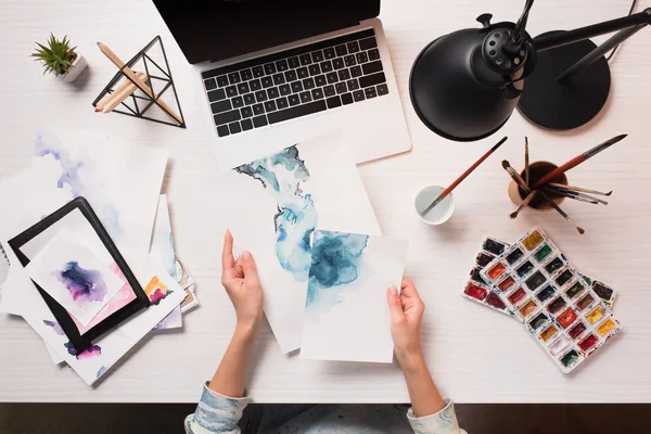 Office desk with laptop, art supplies and cropped view of designer holding drawings, flat lay — Stock Photo