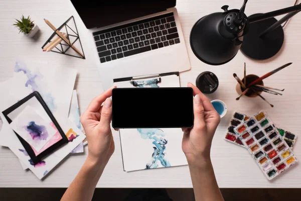 Office desk with laptop, art supplies and cropped view of designer using smartphone with blank screen, flat lay — Stock Photo