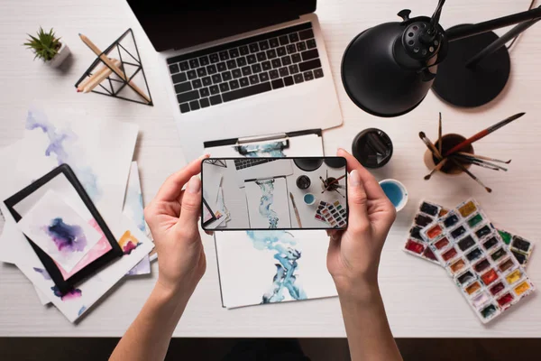 Escrivaninha com laptop, materiais de arte e vista cortada do designer tirando foto com smartphone, flat lay — Fotografia de Stock