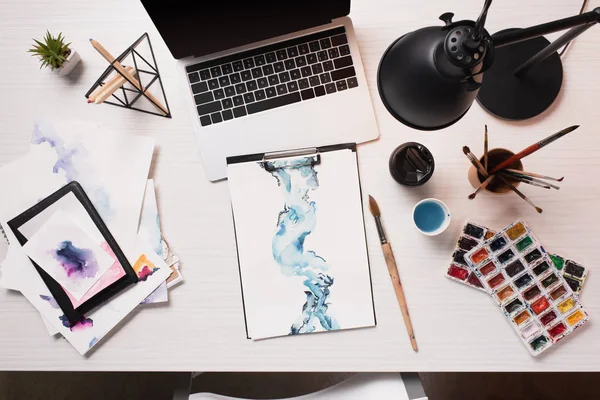 Escritorio de la oficina con el ordenador portátil, bocetos y suministros de arte, disposición plana - foto de stock