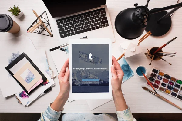 Cropped view of designer holding tablet with tumblr app on screen at office desk with art supplies, flat lay — Stock Photo