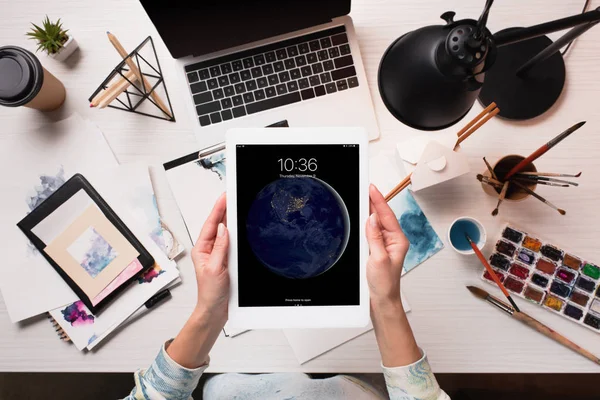 Cropped view of designer holding tablet with apple home screen at office desk with art supplies, flat lay — Stock Photo