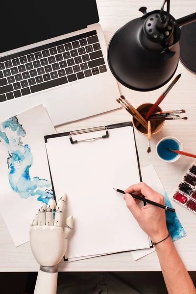 Office desk with laptop, blank sheet and cropped female hand with robot hand, flat lay — Stock Photo