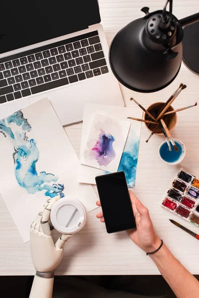 Vista cortada do designer na mesa de escritório com mão robô segurando smartphone com tela em branco — Fotografia de Stock