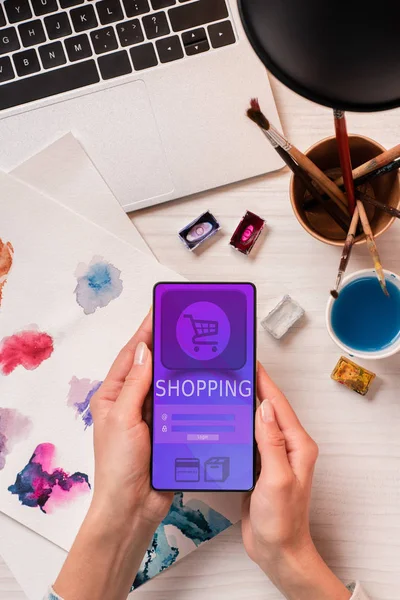 Cropped view of designer at office desk holding smartphone with online shopping app on screen, flat lay — Stock Photo