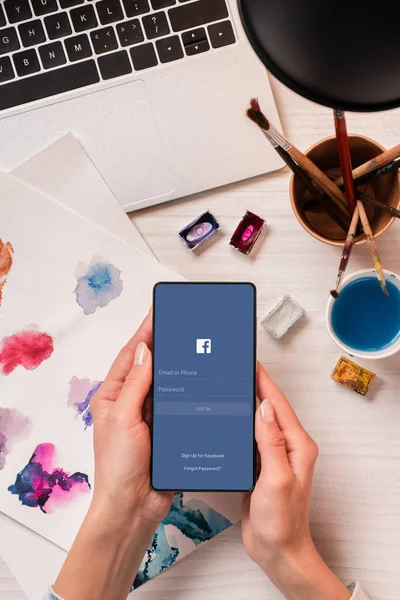 Cropped view of designer at office desk holding smartphone with facebook app on screen, flat lay — Stock Photo