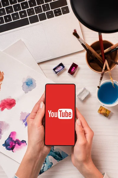 Cropped view of designer at office desk holding smartphone with youtube app on screen, flat lay — Stock Photo