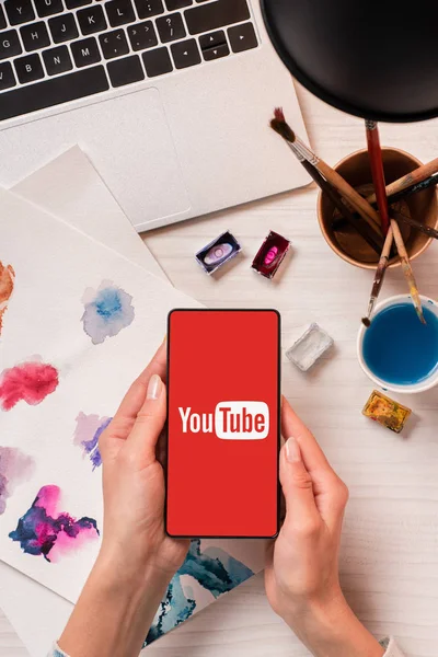 Vista cortada do designer na mesa de escritório segurando smartphone com aplicativo youtube na tela, flat lay — Fotografia de Stock