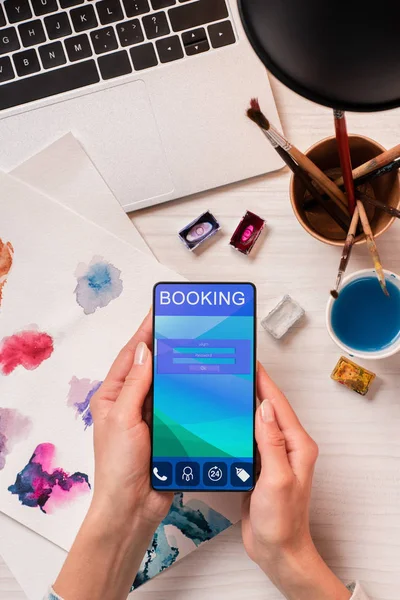 Обрезанный вид дизайнера на рабочем столе со смартфоном с приложением бронирования на экране, плоский лежал — стоковое фото