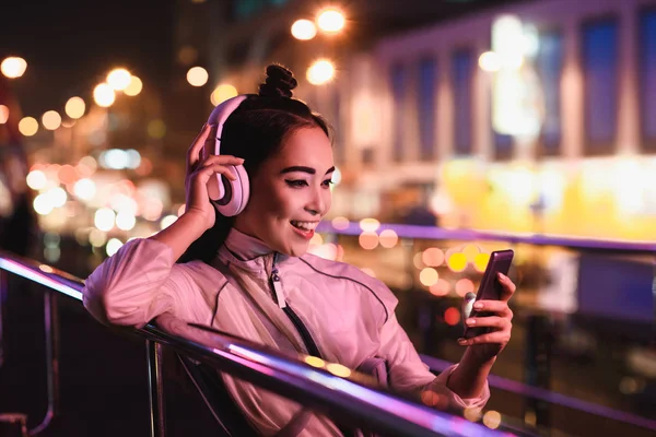 Feliz chica asiática atractiva escuchando música con teléfono inteligente en la calle con luz de neón en la noche, ciudad del concepto futuro - foto de stock