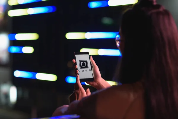 Vue arrière de la fille tenant smartphone avec page uber chargée sur la rue avec lumière au néon dans la soirée, ville du concept futur — Photo de stock