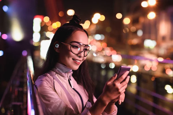 Attractive asian girl with wireless earphone and glasses using smartphone on street with neon light, city of future concept — Stock Photo