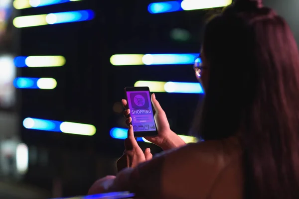 Vista posteriore della ragazza che tiene smartphone con apparecchio per lo shopping in strada con luce al neon in serata, città del concetto futuro — Foto stock