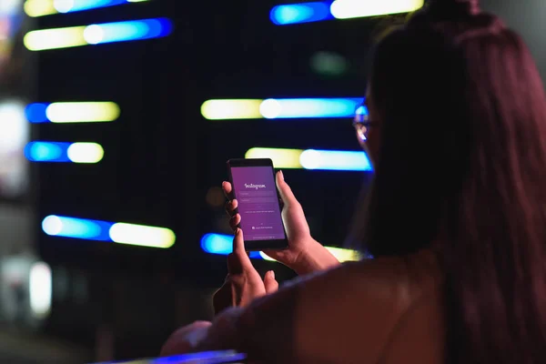 Vue arrière de la fille tenant smartphone avec la page instagram chargé sur la rue avec la lumière au néon dans la soirée, ville du concept futur — Photo de stock