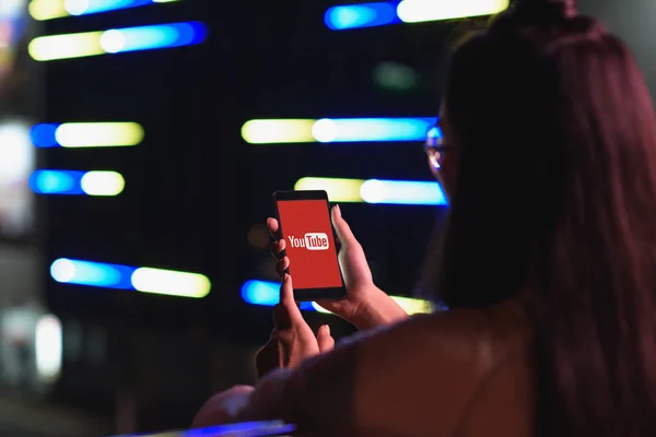 Vue arrière de la fille tenant smartphone avec chargé youtube page sur rue avec néon lumière dans la soirée, ville du concept futur — Photo de stock