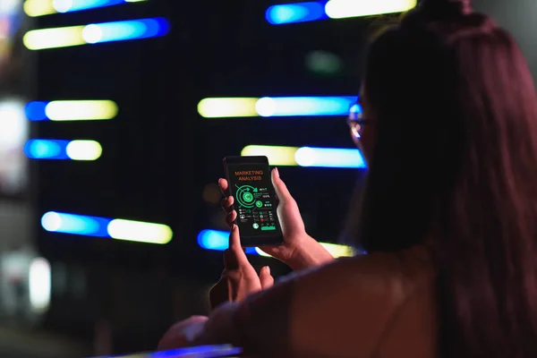 Vista posteriore della ragazza che tiene smartphone con apparecchio di analisi di marketing sulla strada con luce al neon in serata, città del concetto futuro — Foto stock