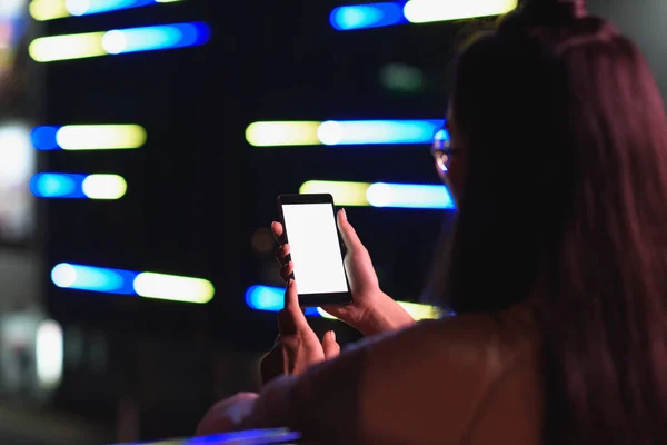 Вид сзади девушки, держащей смартфон с белым экраном на улице с неоновым светом вечером, город будущего — стоковое фото