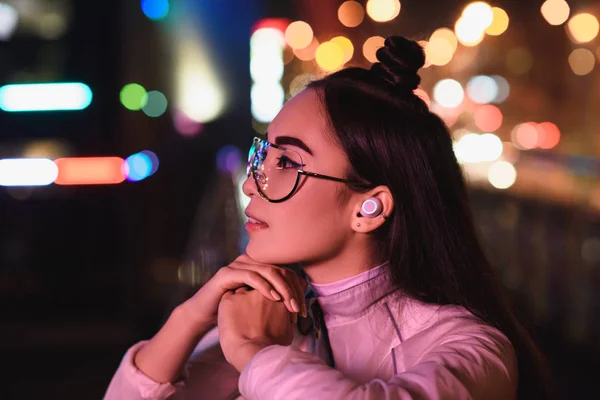 Vue latérale de la fille asiatique élégante dans des lunettes et un écouteur sans fil regardant loin sur la rue avec lumière au néon le soir, ville du concept futur — Photo de stock