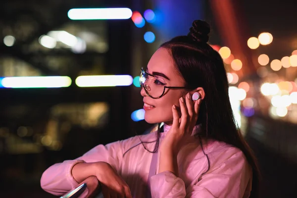 Stylish asian girl in glasses touching wireless earphone on street with neon light in evening, city of future concept — Stock Photo