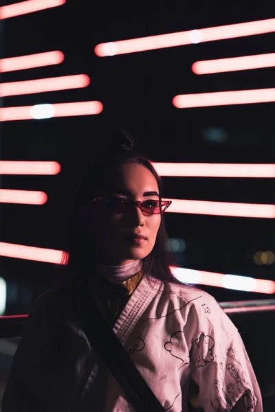 Attraktive asiatische Mädchen in weißem Kimono und Sonnenbrille, die abends mit Neonlicht auf der Straße stehen, Stadt der Zukunft — Stockfoto