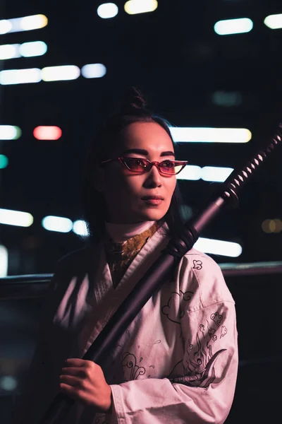 Atractiva chica asiática en kimono blanco sosteniendo espada en la calle con luz de neón en la noche, ciudad del concepto futuro - foto de stock