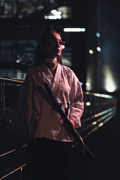 Attractive asian girl in white kimono holding katana on street with neon light in evening, city of future concept — Stock Photo