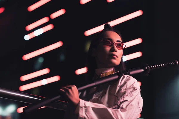 Niedrigwinkel-Ansicht attraktiver asiatischer Mädchen im weißen Kimono mit japanischem Schwert auf der Straße mit Neonlicht am Abend, Stadt der Zukunft Konzept — Stockfoto