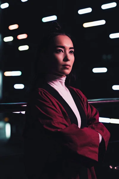 Приваблива азіатська дівчина в бургундському кімоно, що стоїть з схрещеними руками на вулиці з неоновим світлом ввечері, місто майбутньої концепції — стокове фото