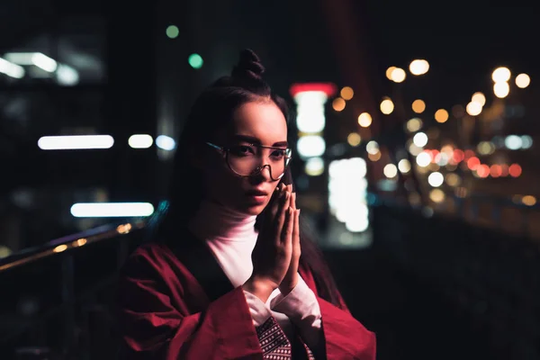 Beautiful asian girl in burgundy kimono making namaste gesture on street with neon light in evening, city of future concept — Stock Photo