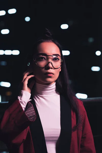 Atractiva chica asiática en kimono de color burdeos y auriculares inalámbricos de pie en la calle con luz de neón en la noche, ciudad del concepto futuro - foto de stock