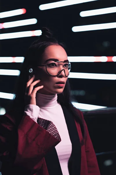 Atractiva chica asiática en kimono de color burdeos y auriculares inalámbricos mirando hacia otro lado en la calle con luz de neón en la noche, ciudad del concepto futuro - foto de stock