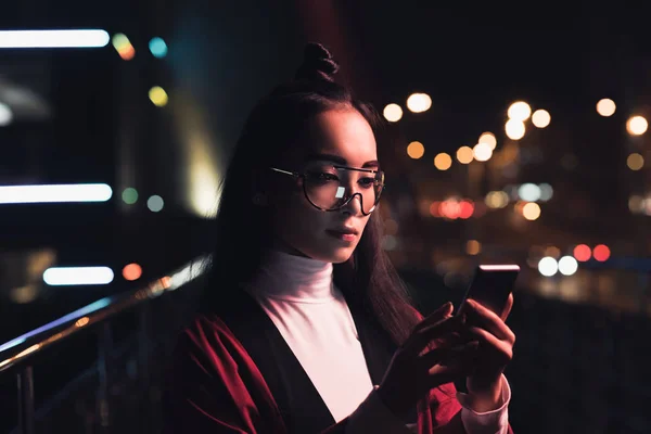 Приваблива азіатська дівчина в бургундському кімоно і окулярах, використовуючи смартфон на вулиці з неоновим світлом ввечері, місто майбутньої концепції — стокове фото